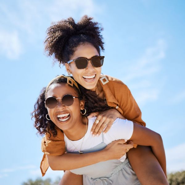 Summer, friends and freedom with women and piggy back against the blue sky for hug, lifestyle and happy on Puerto rico holiday. Youth, crazy and friend with girl on vacation with glasses and a smile.